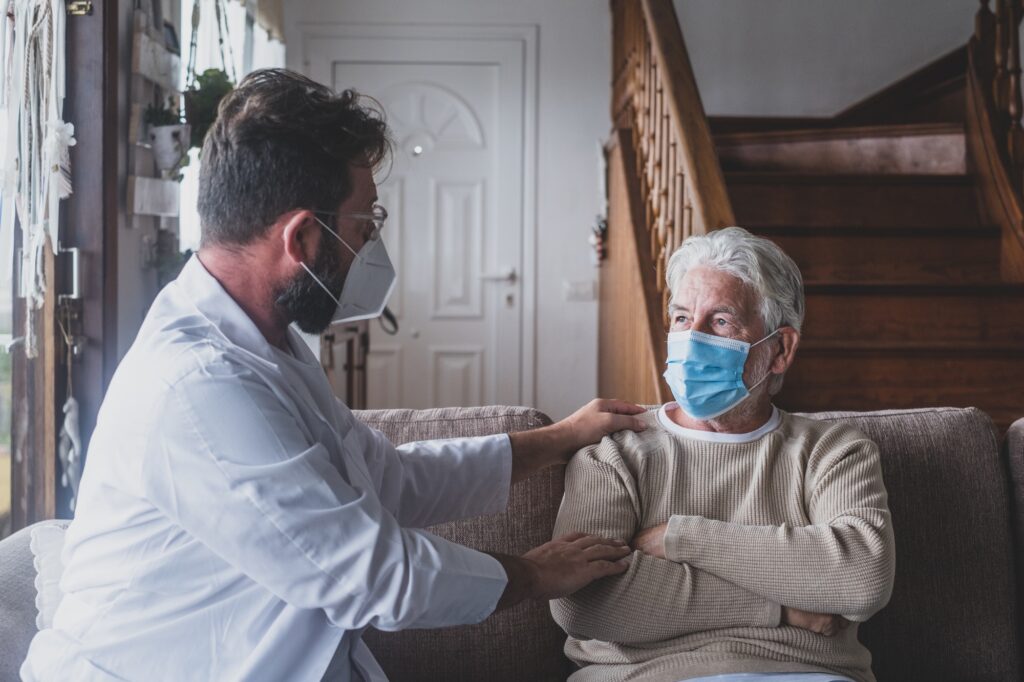 Male professional doctor consulting senior patient during medical care visit wearing masks.