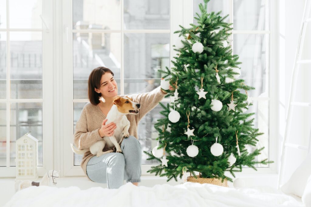 Lovely happy woman holds pedigree dog on hands, decorates New Year tree