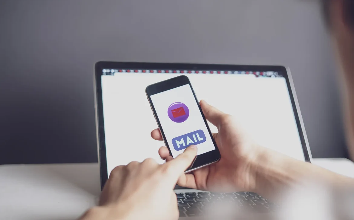 Bir kişi, mobil cihazında e-posta uygulamasını açarken arka planda bir dizüstü bilgisayar ekranı görünüyor.